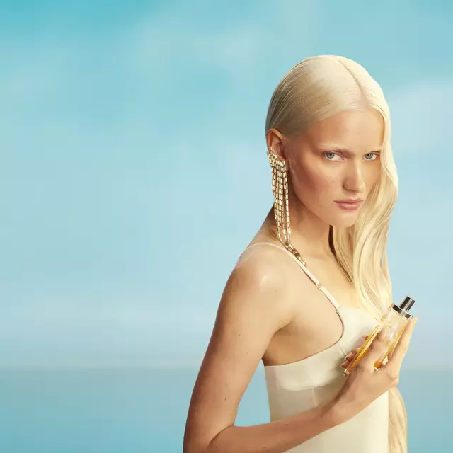 New Mugler Starlicious les eaux gourmandes campaign banner featuring a model with long blonde hair and blue eyes, wearing a pale yellow dress, holding the Pineapple Musk fragrance bottle against a soft blue sky background. The fragrances are delicious, collectable and layerable.